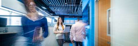 Woman holding laptop stands in focus in modern office while motion-blurred coworkers move around her, creating a dynamic workplace scene with contemporary blue and wood interior design.