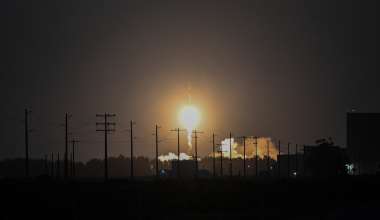 Nighttime rocket launch viewed from a distance, with a bright flame and exhaust illuminating the horizon. Utility poles and power lines are silhouetted in the foreground against the dark sky. 