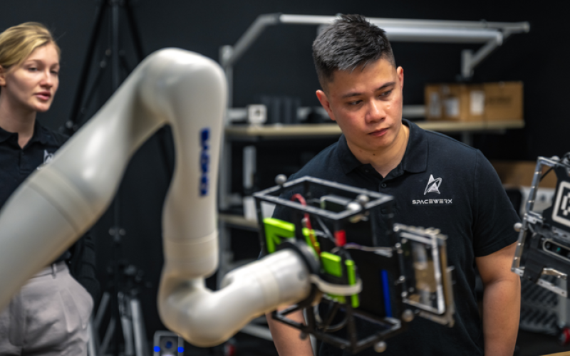 A man and a woman stand in a dark laboratory observing equipment. In front of them is a white robotic arm. The man's t-shirt has a logo for SpaceWERX.