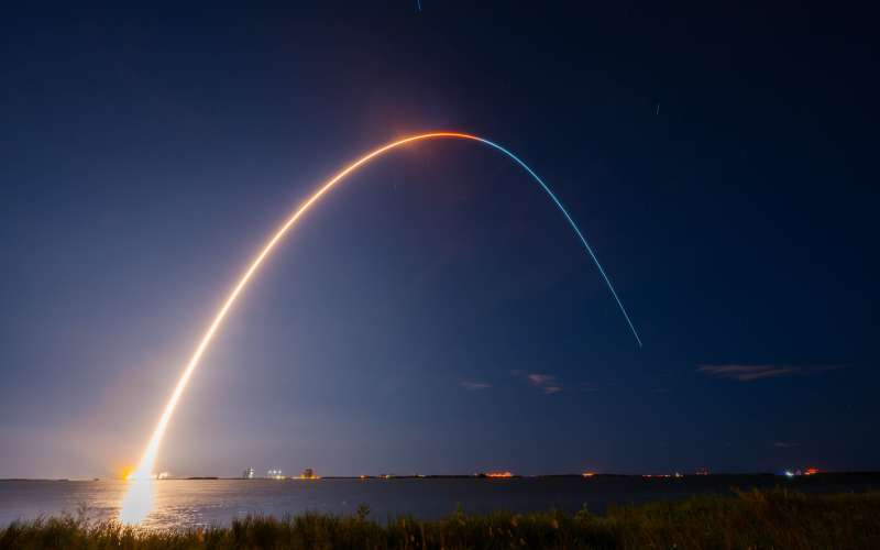 The image captures a nighttime rocket launch. The scene features a long-exposure photograph that shows the bright, curved trail of the rocket as it ascends into the dark sky. The trail starts from the brightly lit launch pad on the horizon near the left side of the image and arcs gracefully across the sky, fading towards the right. The sky is mostly clear, with a few scattered clouds and faint stars visible. In the foreground, there is a grassy area leading to a body of water that reflects the light.