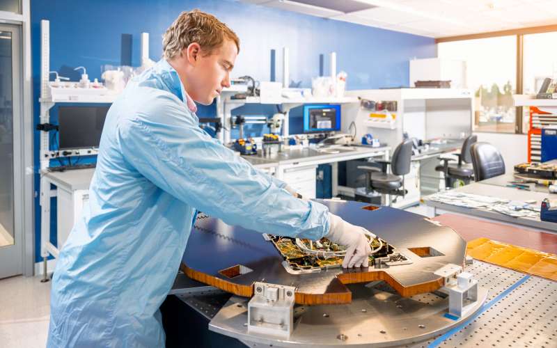 A person in a blue lab coat and white gloves is working in a high-tech laboratory or cleanroom. They are handling a large flat disk-shaped satellite on a specialized table. The background features organized workstations with monitors, tools, and other electronic devices, indicating a professional and controlled environment. The room is brightly lit with natural light streaming through large windows.