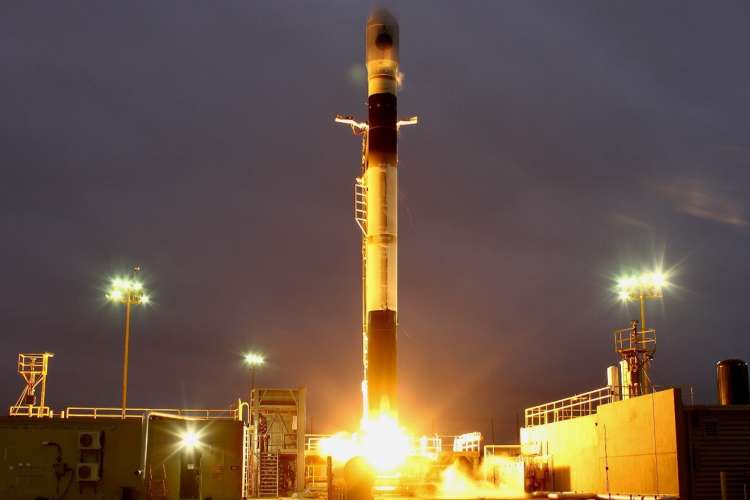 The image shows a rocket launching into the sky at night. The rocket is standing vertically on a launch pad, with bright flames and exhaust gases emitting from its base, illuminating the surrounding area. The rocket itself is tall, cylindrical, and mostly white with a black section near the top. The launch pad is equipped with various structures and equipment, and several floodlights are seen around the area, casting a bright light against the dark, cloudy sky. 