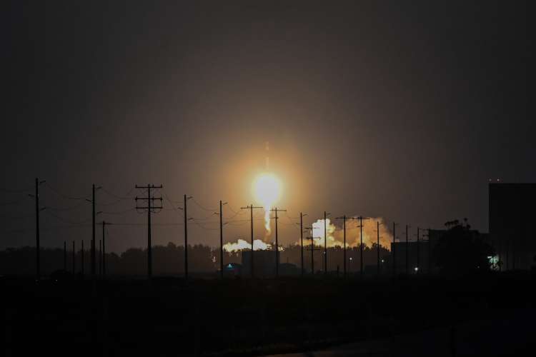 Nighttime rocket launch viewed from a distance, with a bright flame and exhaust illuminating the horizon. Utility poles and power lines are silhouetted in the foreground against the dark sky. 