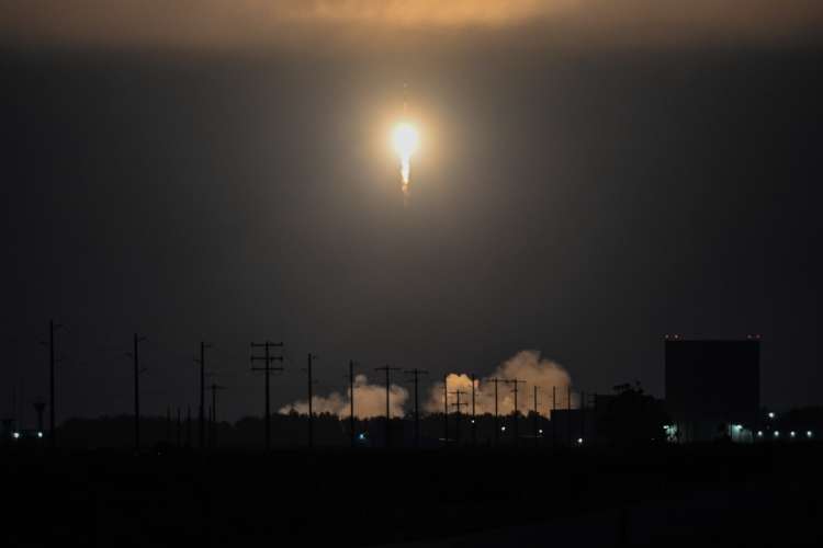 Nighttime rocket launch viewed from a distance, with a bright flame and exhaust illuminating the horizon. Utility poles and power lines are silhouetted in the foreground against the dark sky.