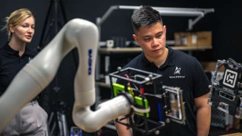 A man and a woman stand in a dark laboratory observing equipment. In front of them is a white robotic arm. The man's t-shirt has a logo for SpaceWERX.