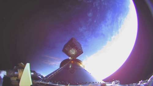 The image captures a view from an onboard camera of a satellite being deployed into space. In the foreground, parts of the deploying mechanism and the satellite itself are visible. The satellite appears to be a cube-shaped object mounted on top of a conical structure. In the background, the curvature of Earth is prominently displayed, with the planet's surface partially illuminated by sunlight, creating a striking contrast between the lit and shadowed areas of the Earth.