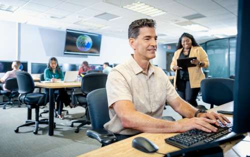 Man with woman in the background looking at a computer monitor.