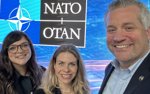 Two women and a man posing in front of the NATO logo.